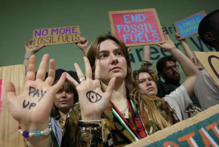 A group of protesters stand holding signs, they have eyes drawn on their palms