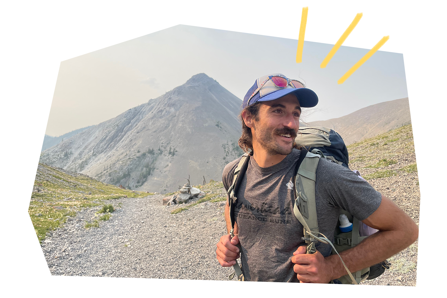 Jeff, wearing backpack and hat, looking off in distance in front of mountain landscape