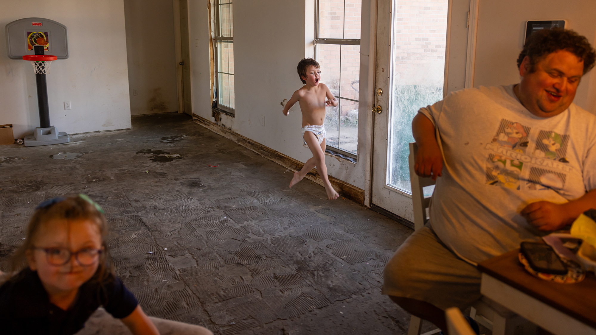 A middle-aged man sits at a table in a house with severely damaged floors and walls. Nearby, two children can be seen. One is lit by a warm indoor light and slightly out of focus. The other is running through the house wearing only a diaper.