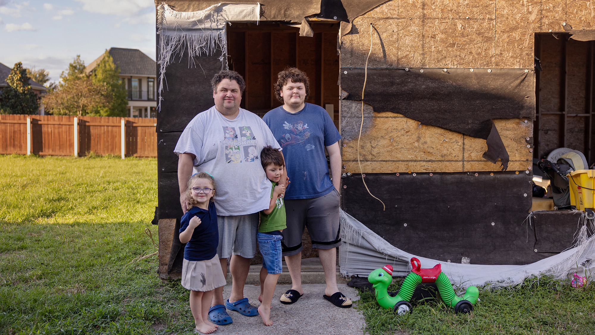 A man and three children -- one boy as tall as the man and two younger children hugging the man -- stand in front of a severely damaged house with plywood boards and torn mesh tarp. The lawn outside is green and tehre is a plastic toy worm with a saddle visible to the side of the walkway.