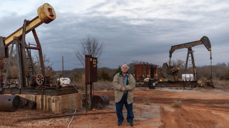 A man stands outdoors with oil drills to either side of him. He has white hair and his hands are clasped in front of him.