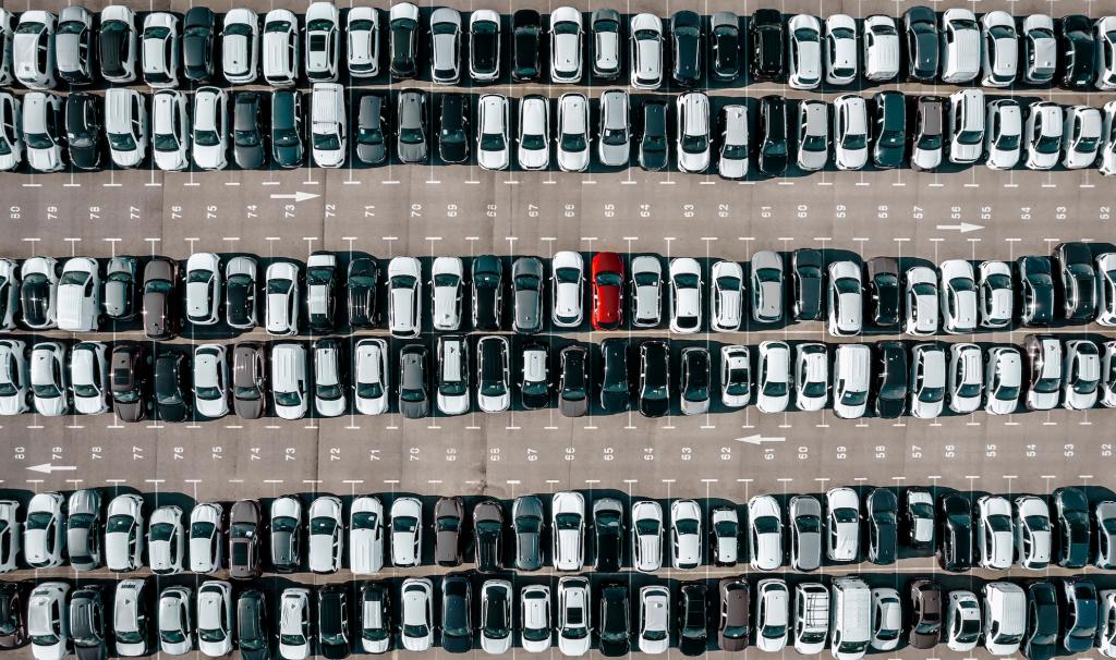 An aerial view of a vast parking lot filled with rows of black, white, and gray cars, creating a uniform pattern. Among them, a single red car stands out, drawing attention with its bold contrast.
