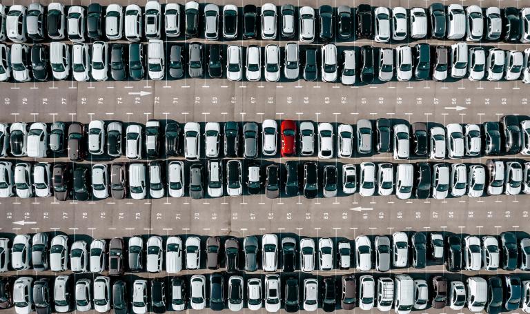 An aerial view of a vast parking lot filled with rows of black, white, and gray cars, creating a uniform pattern. Among them, a single red car stands out, drawing attention with its bold contrast.