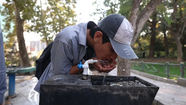 A man in a baseball cap cups water in his hand from a water fountain