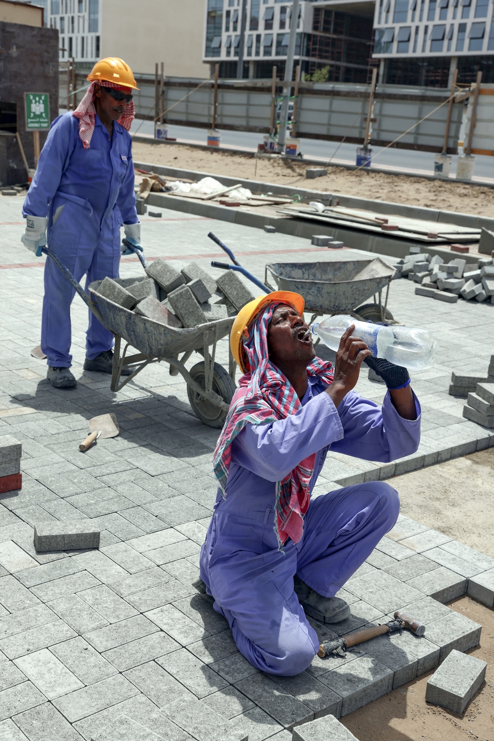 A man drinks water on a construction site while another pushes a wheelbarrow full of materials