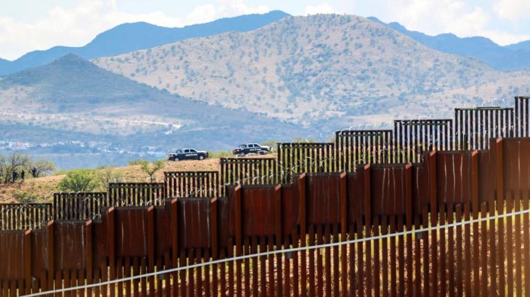 A photo shows a border fence with vehicles in the background.