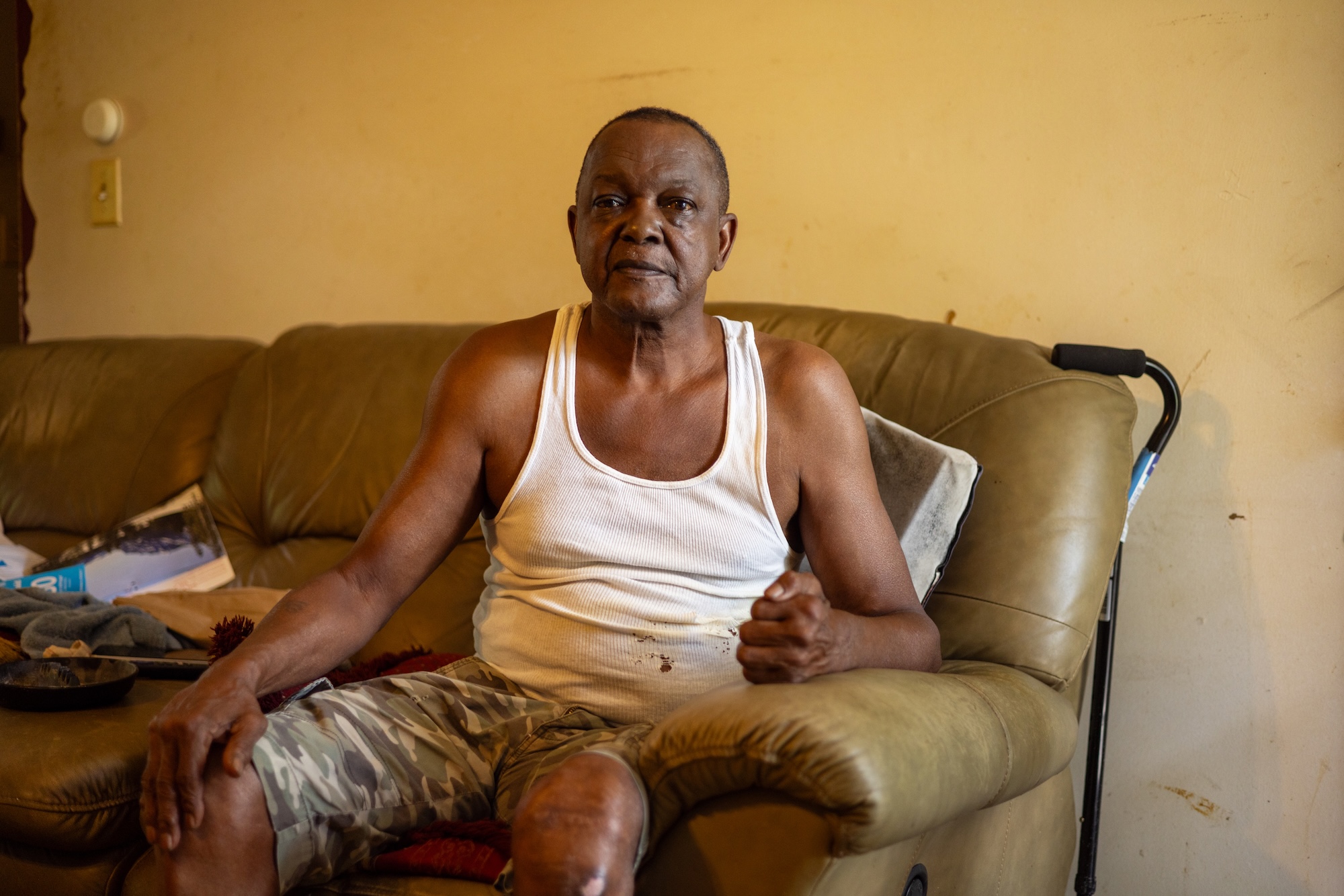 A man in a tank top sits on a brown couch with a cane nearby