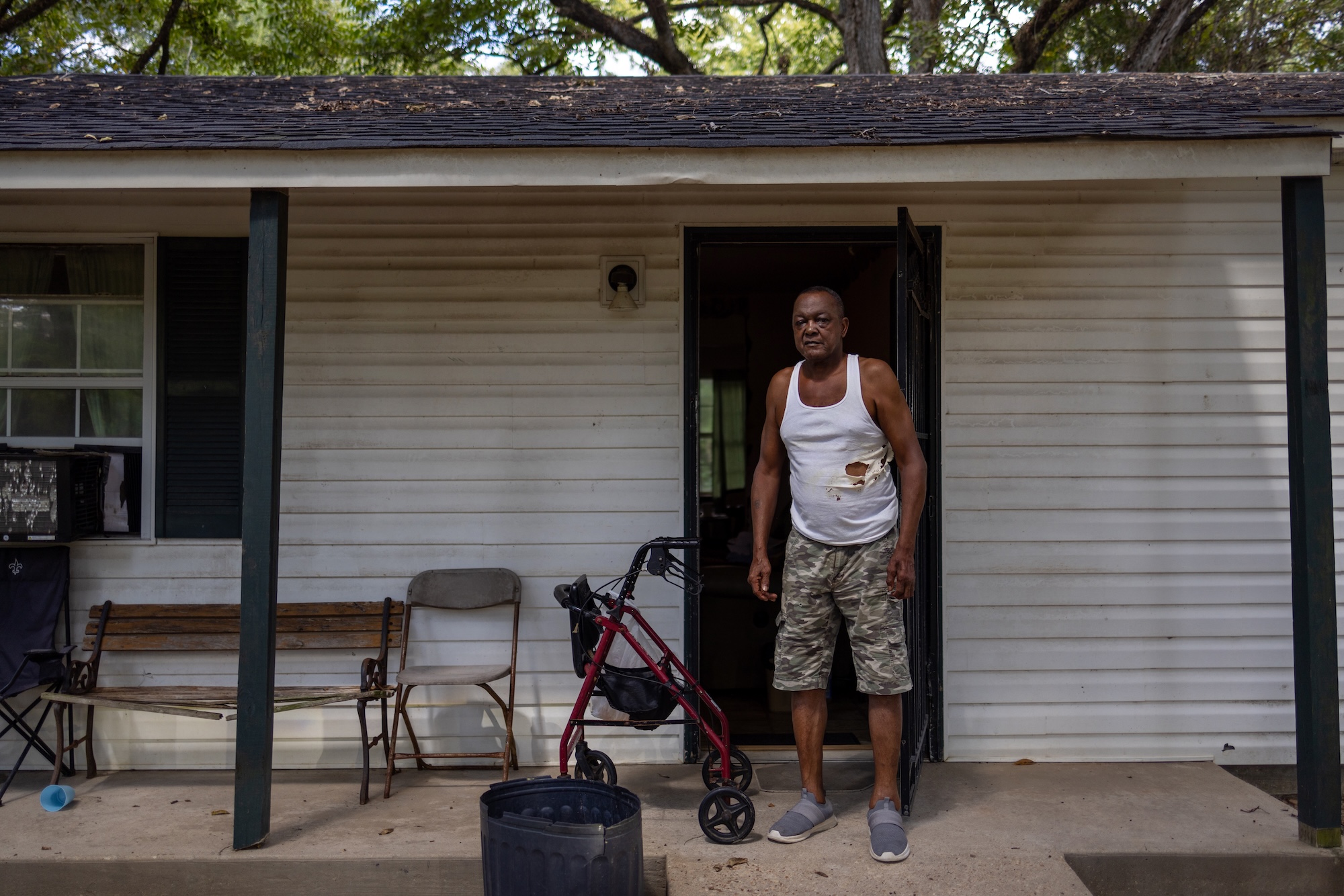 A man in a torn undershirt and shorts stands next to a walker on the porch of his house