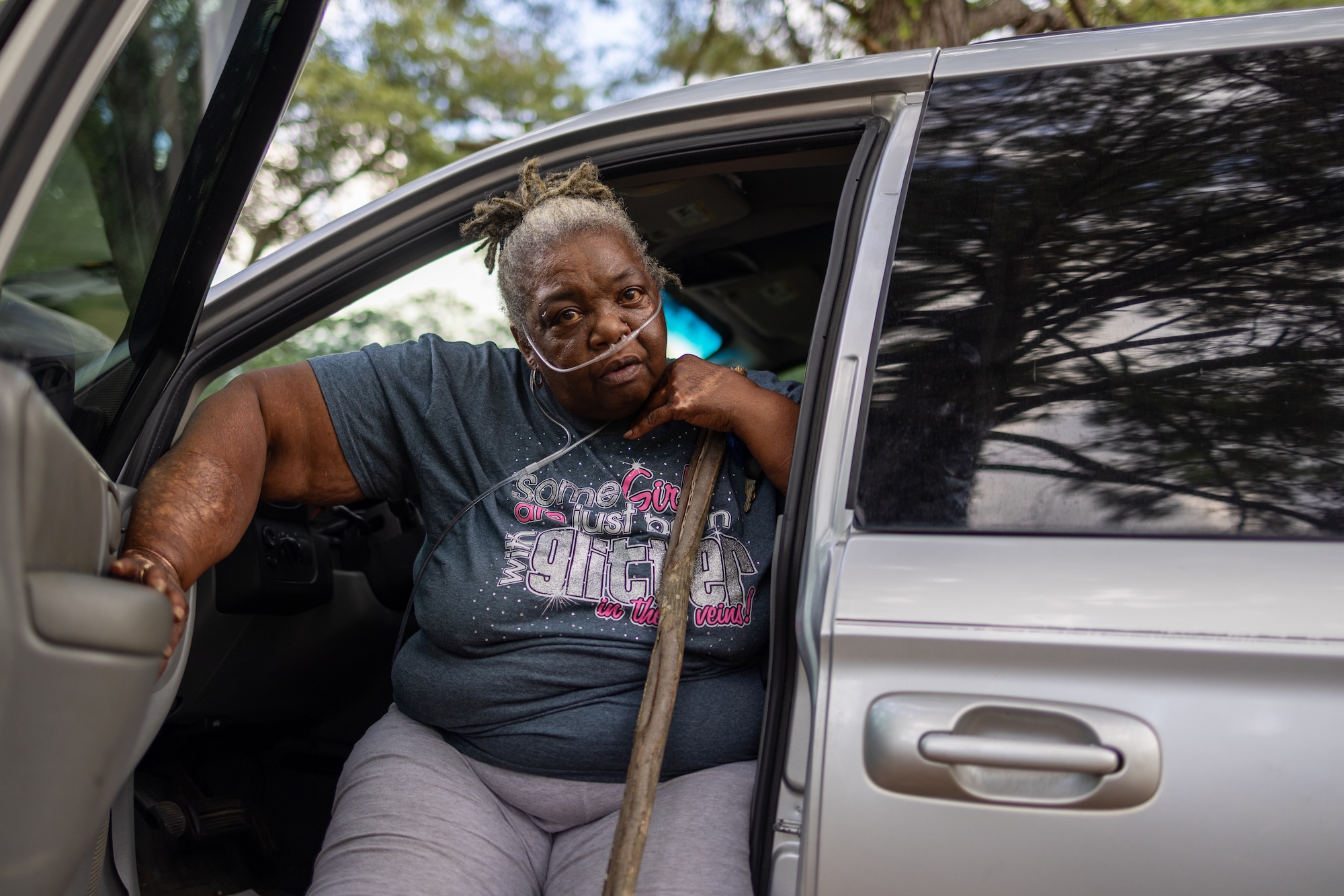A woman sits in a car with the door open. She leans on a cane and wears a breathing apparatus