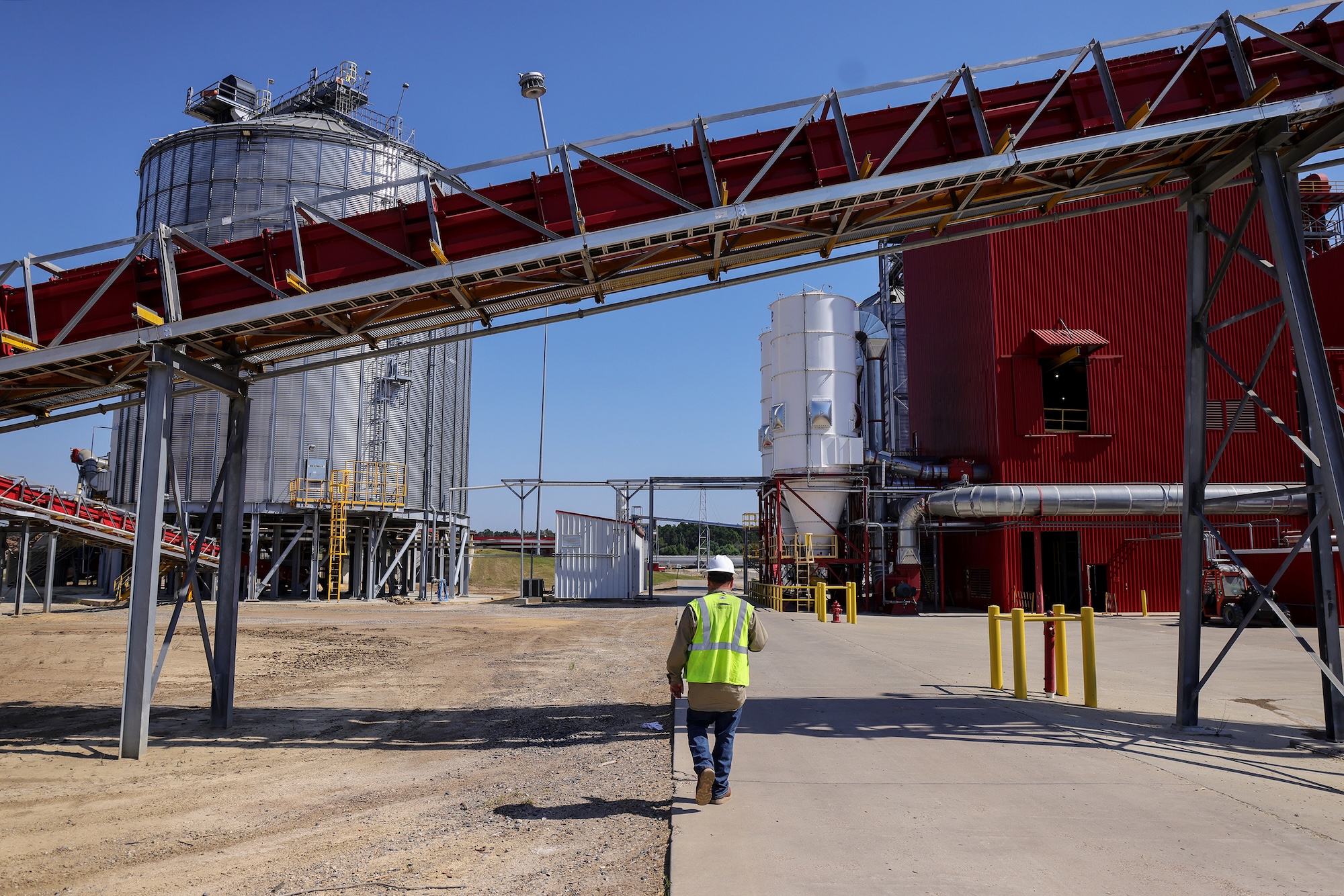 A worker in a neon vest and hard hat walks toward a large storage tank at a biomass facility