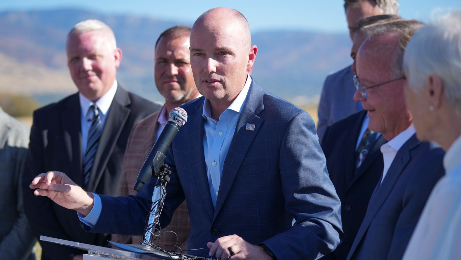 Men in suits stand near the shores of a lake with mountains in the background.