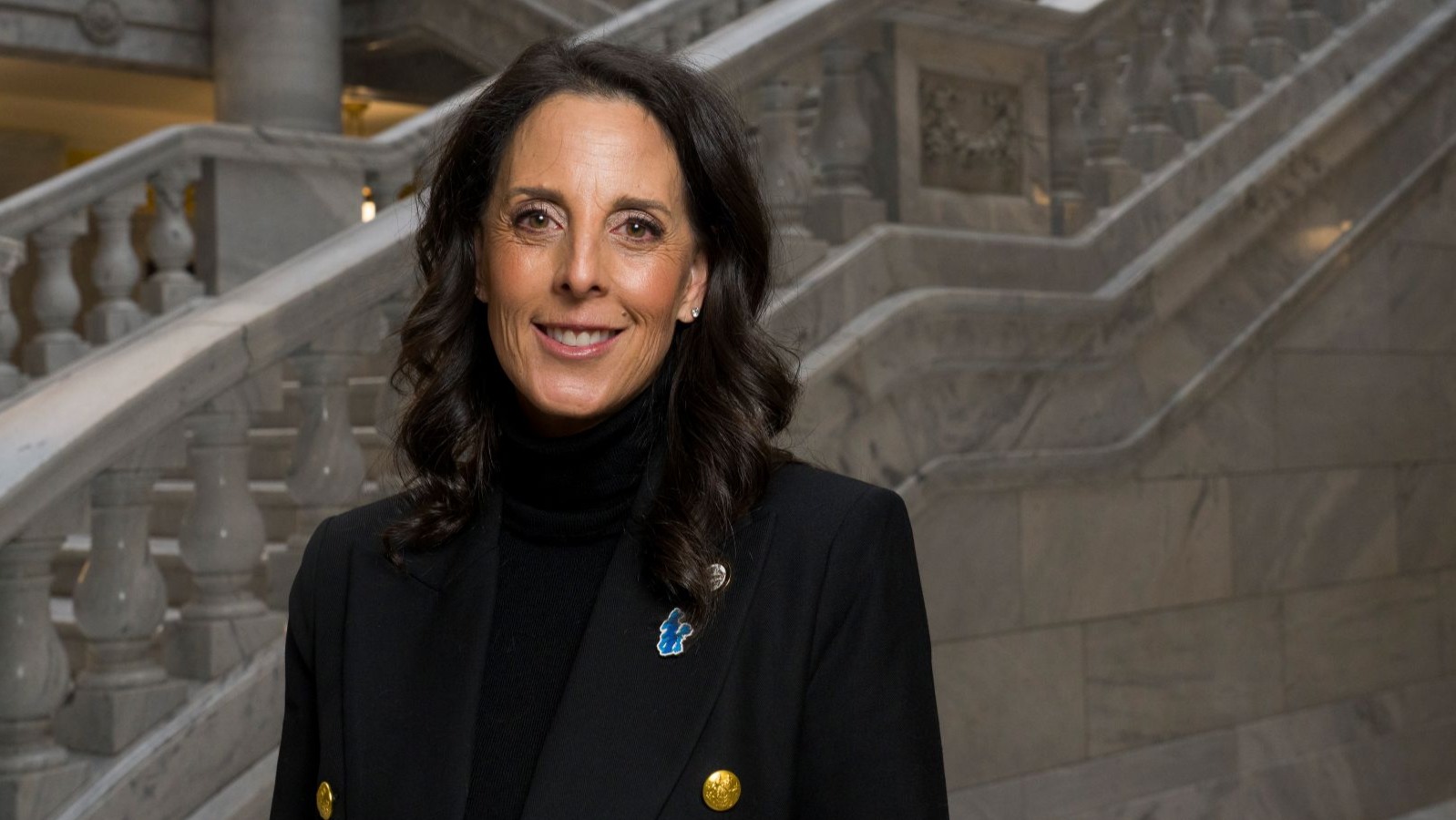 A woman with a black turtleneck, blazer, and pin of the Great Salt Lake stands in front of marble stairs.