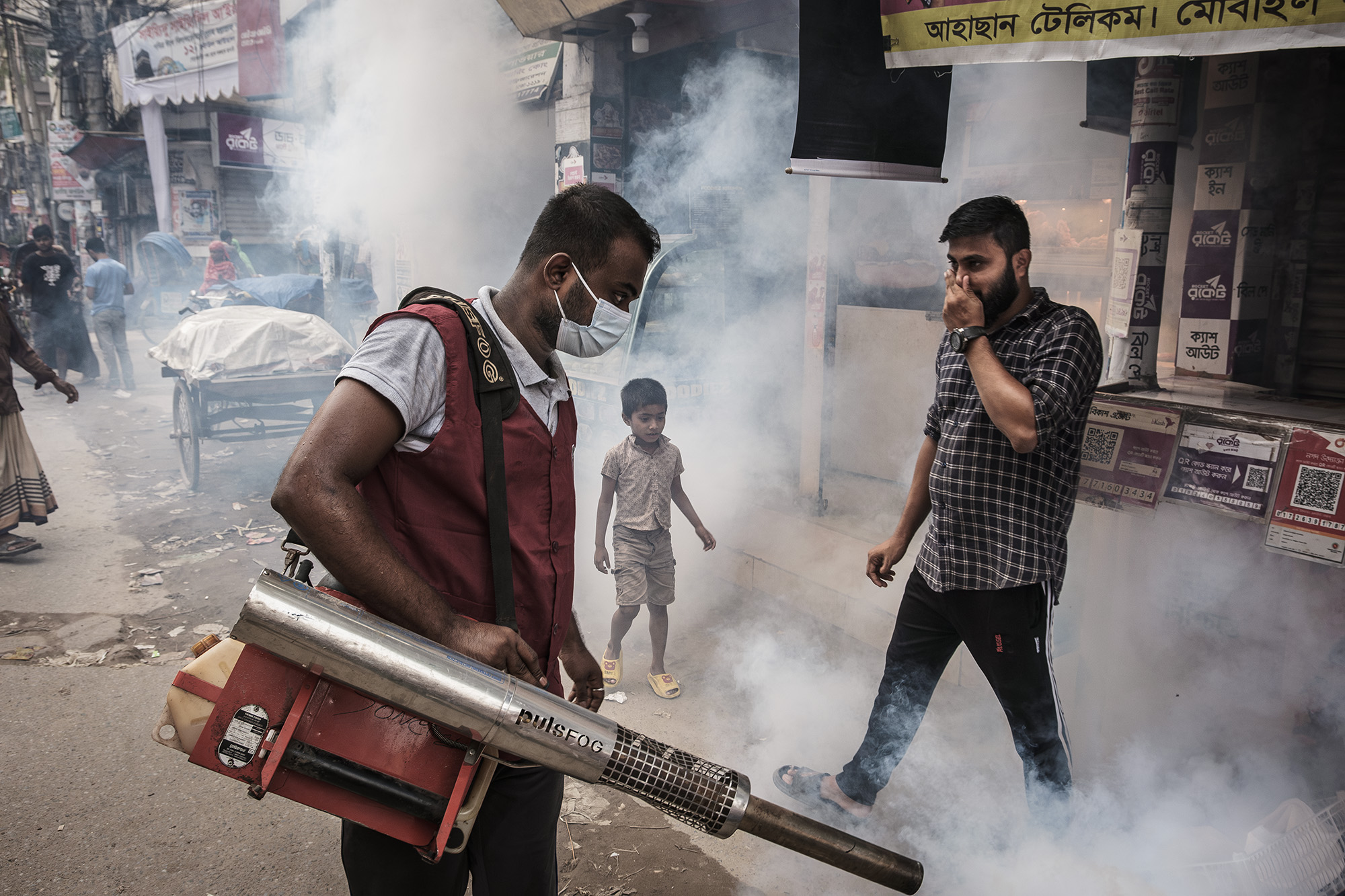 A man fumigates an outdoor stretch of shops while an unmasked adult and child stand nearby