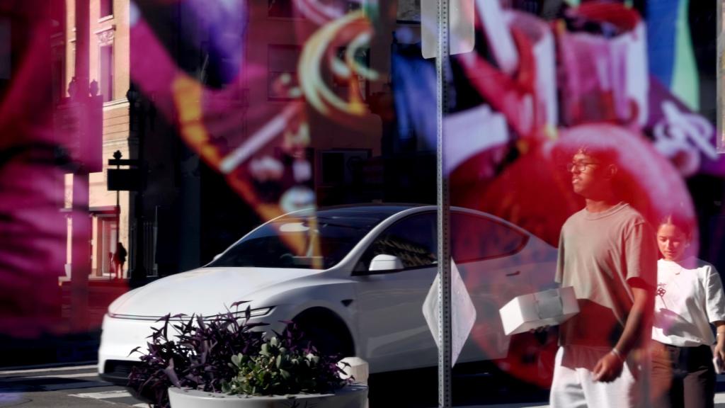 A Tesla car and pedestrians are shown in the reflection of a storefront window in downtown Los Angeles.