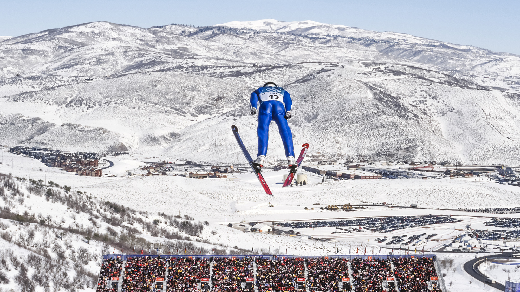 A skiier in a blue skintight suit launches off a giant jump nestled between snowy mountains