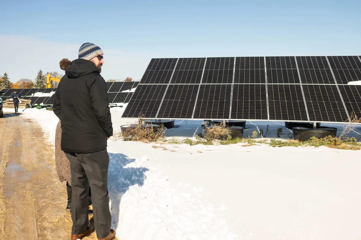 A man in winter clothes stands on the snow next to solar panels