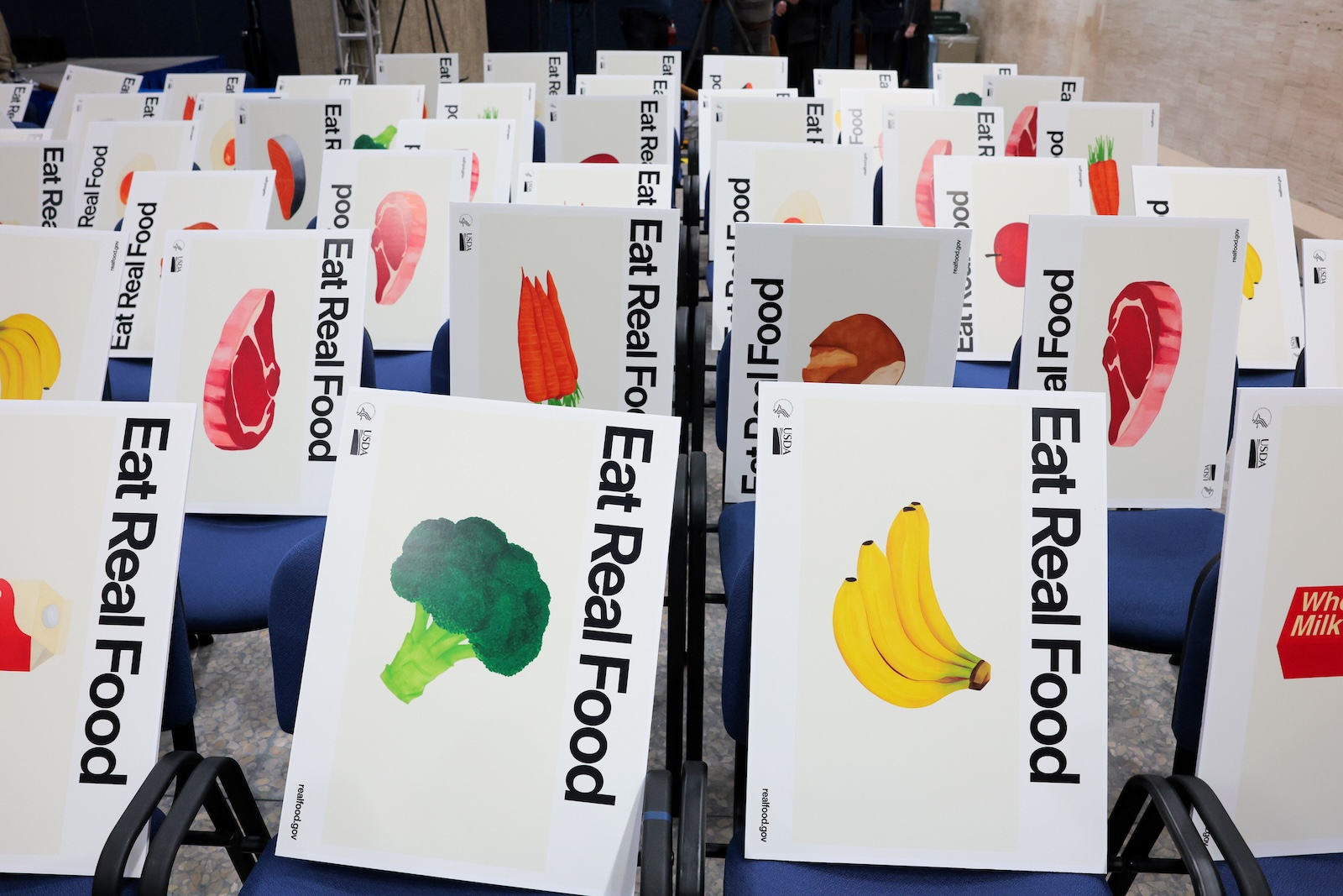 rows of blue chairs in an empty conference room; each chair has a white posterboard resting on it with an image of a fruit, vegetable, or cut of meat with the words "Eat Real Food"