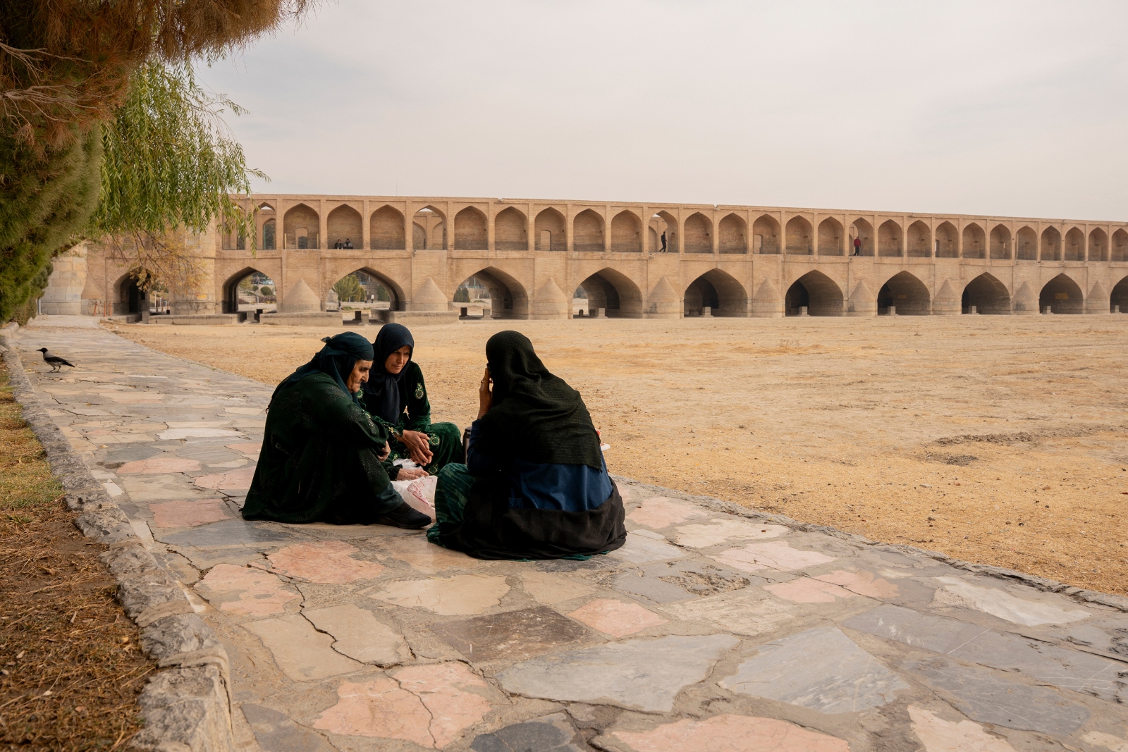 Three Iranian women sit in a group talking on a stone sidewalk along the dried-up riverbed