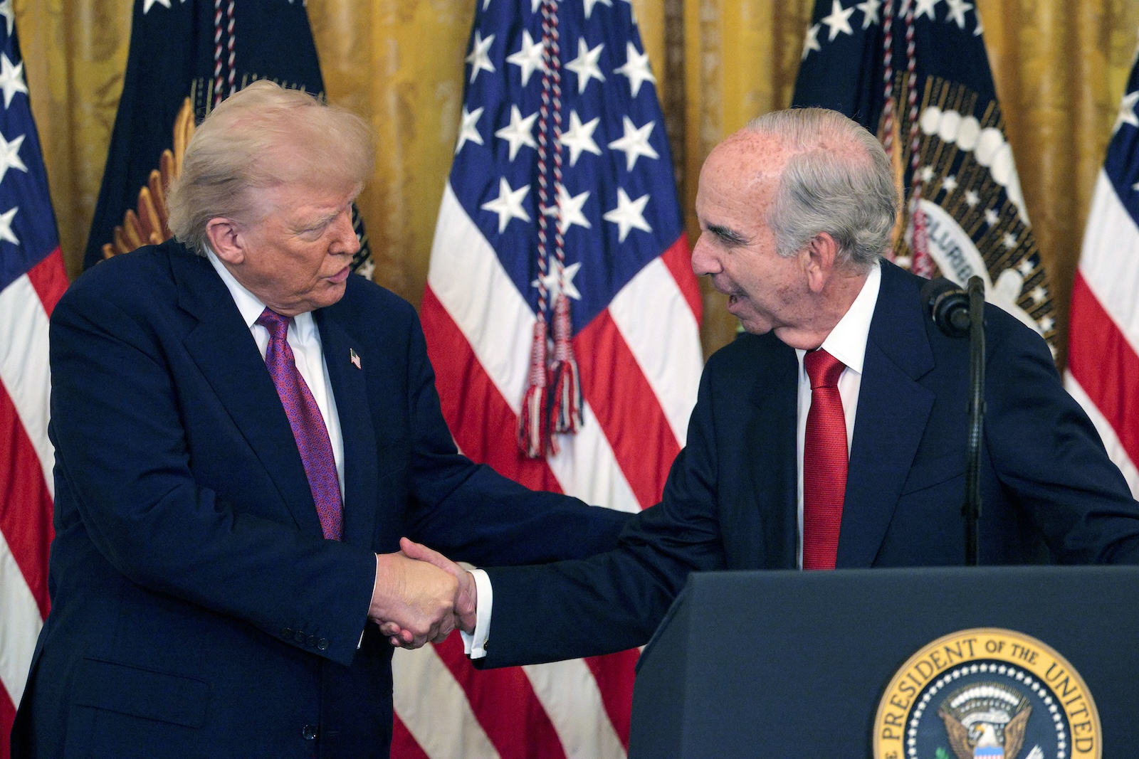 two old men shake hands in front of a row of US flags and behind a podium
