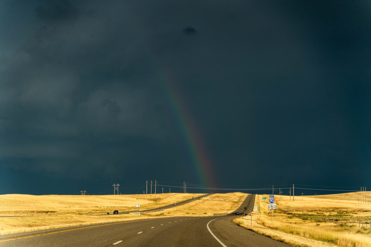 A rainbow stretches over a sunlit prairie and road cutting through the yellow grass