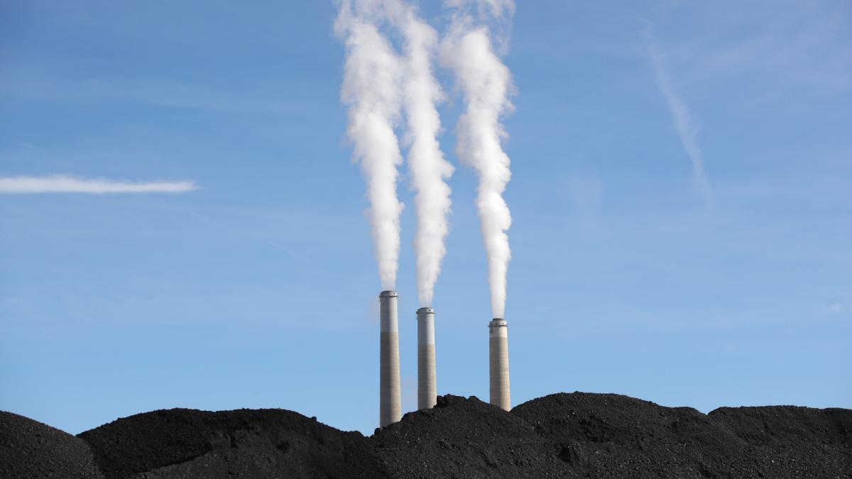 Photo of smokestacks spewing smoke into a blue sky with large mounds of coal in front