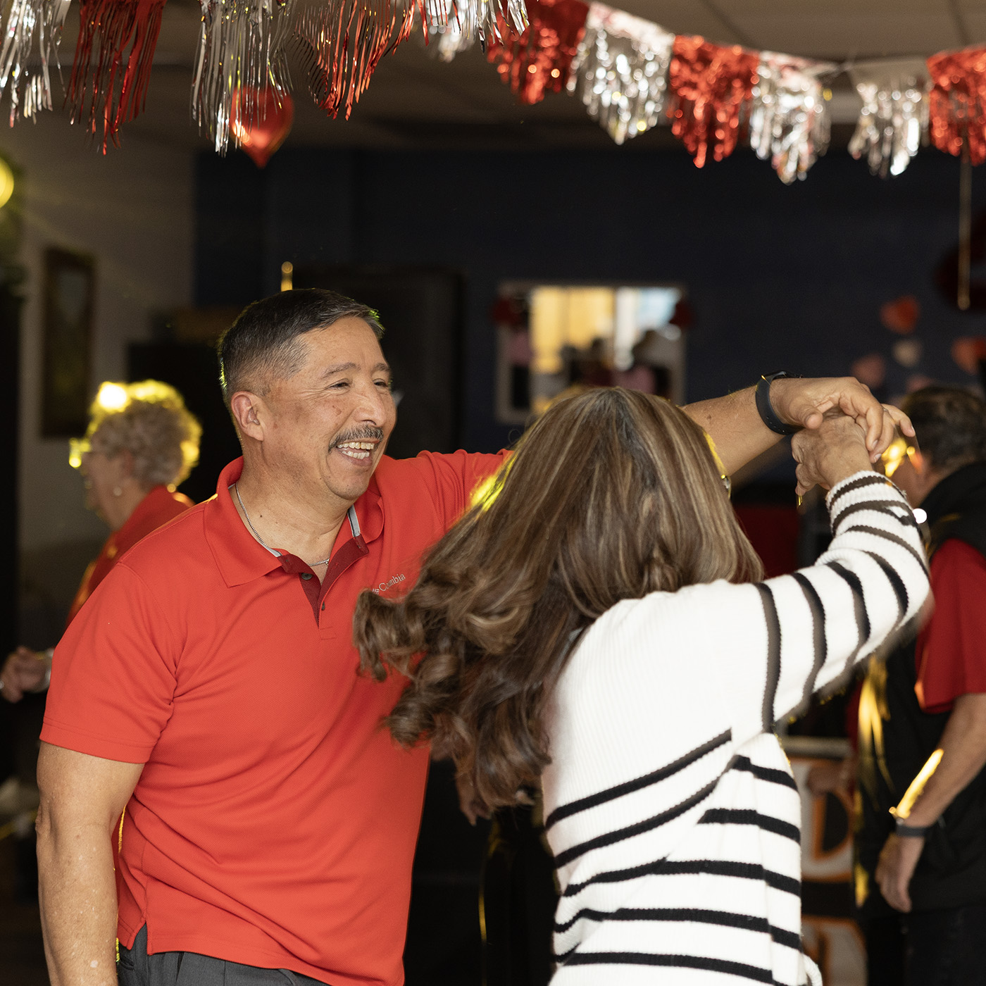 A smiling man in a red shirt twirls a woman on the dance floor under strings of red and white decorations