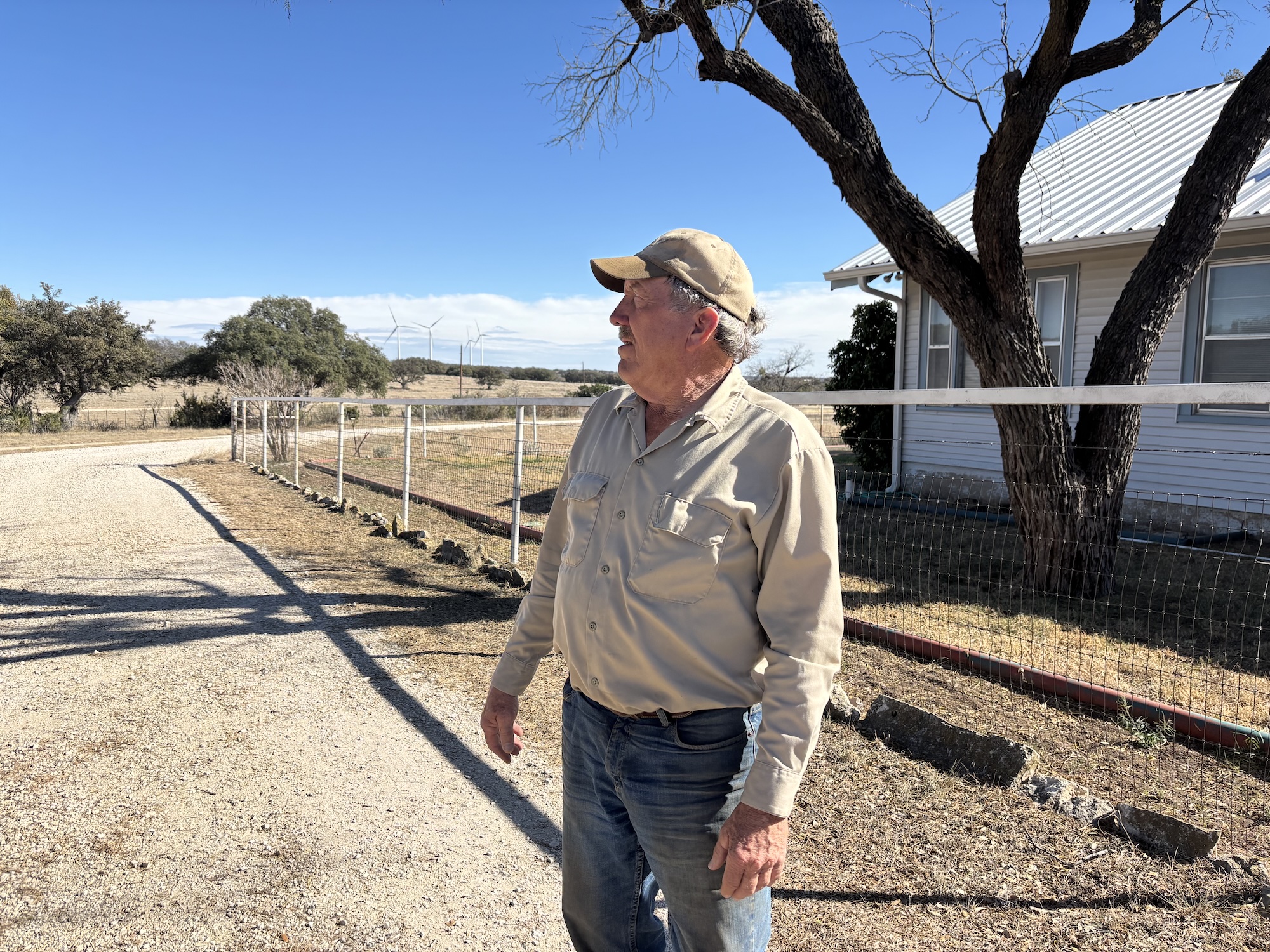 A man in a baseball cap and button up shirt stands on a rural road