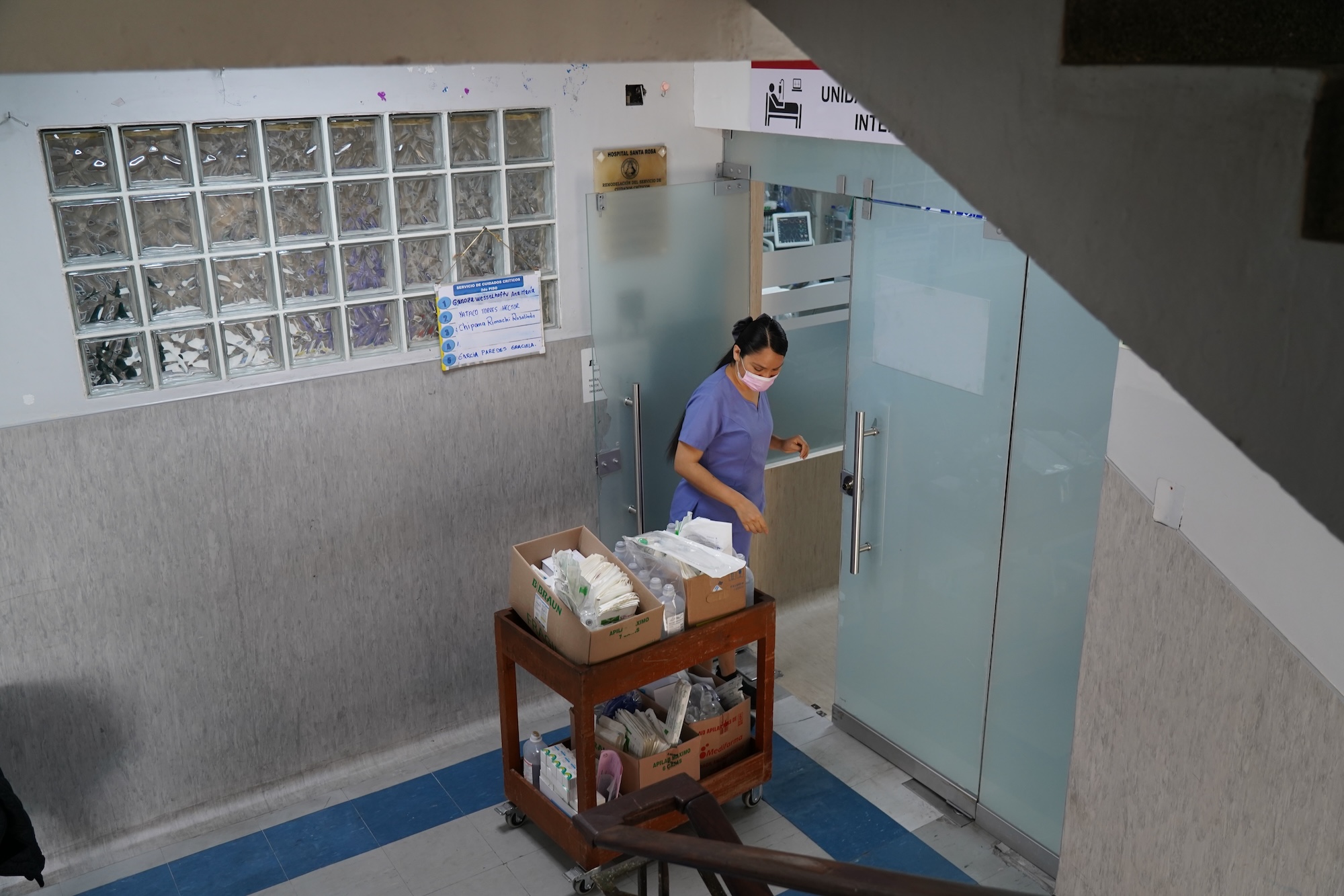 A woman wheels a cart with supplies to a door in a hospital