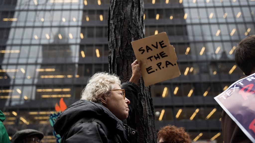 An older woman walks next to an office building holding a Save the EPA handmade sign