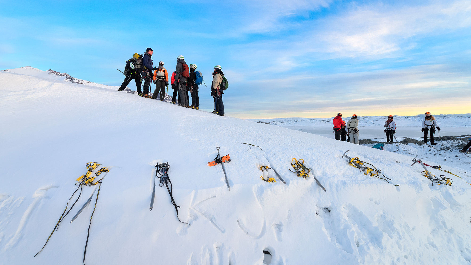 A group of tourists stands on top of a snowy slope, with their crampons lined up in the snow in the foreground