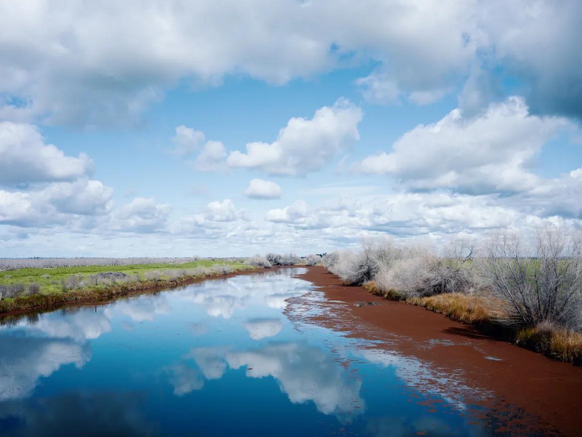 A canal of water flows through a field and reflects clouds