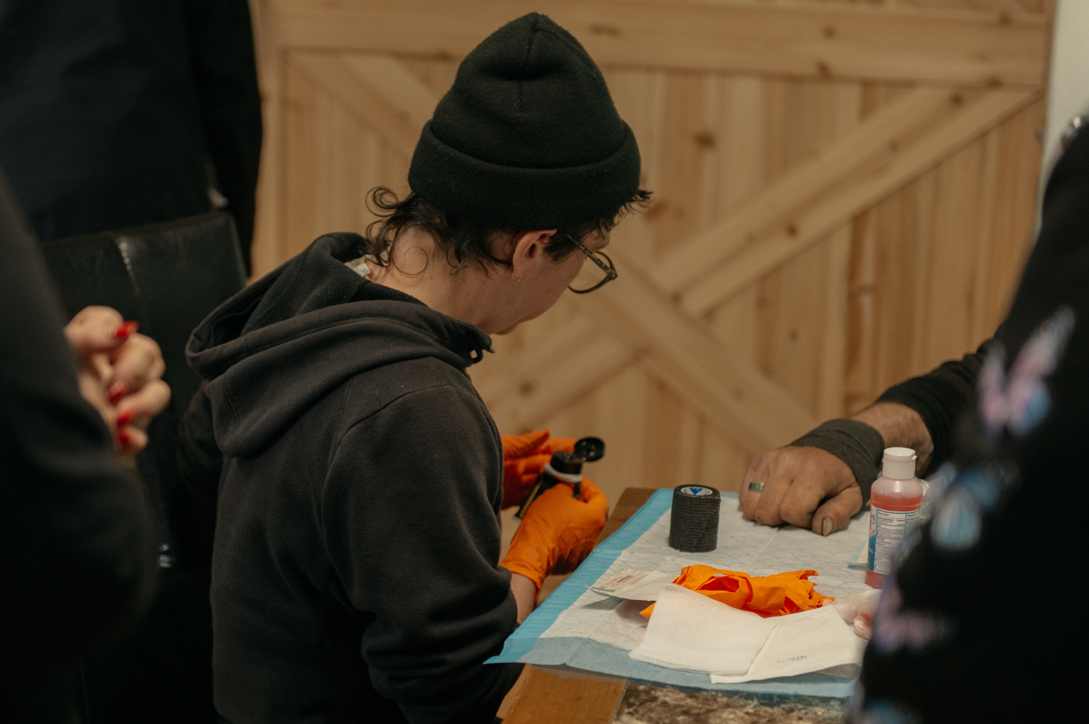 A medical professional, wearing orange latex gloves, prepares ointment, gauze, and a bandage to treat a wound of a patient sitting across the table.