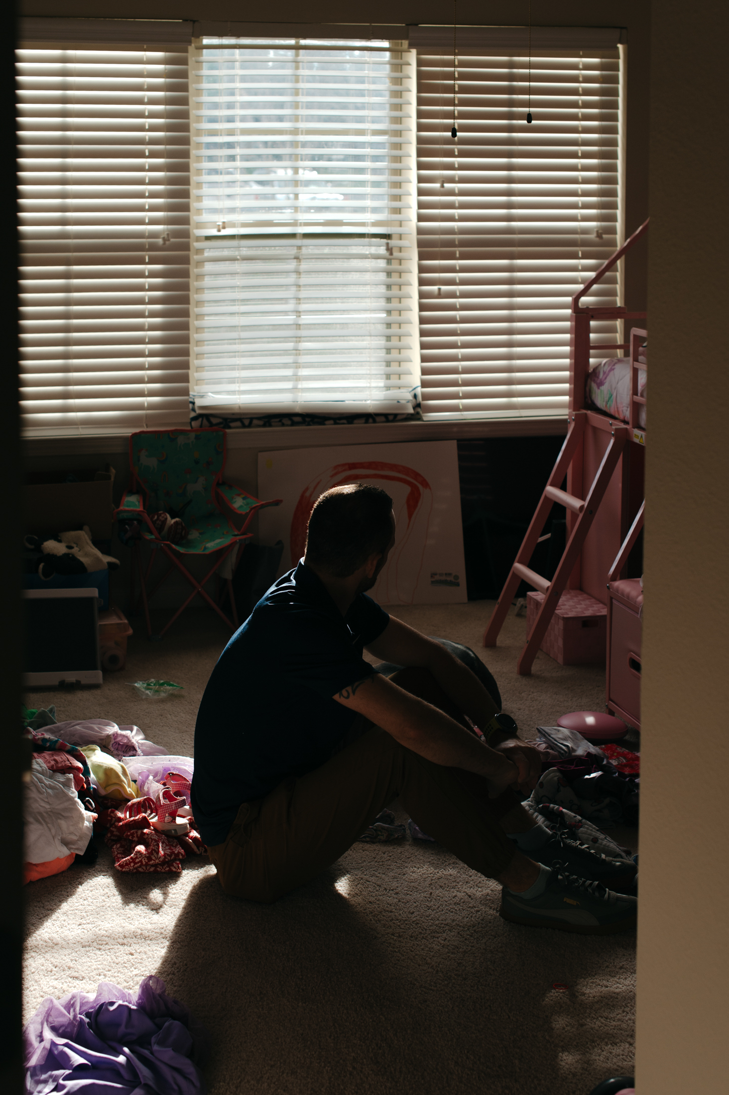 A man sits on the floor of a dimly lit child's bedroom, looking out a bright window.