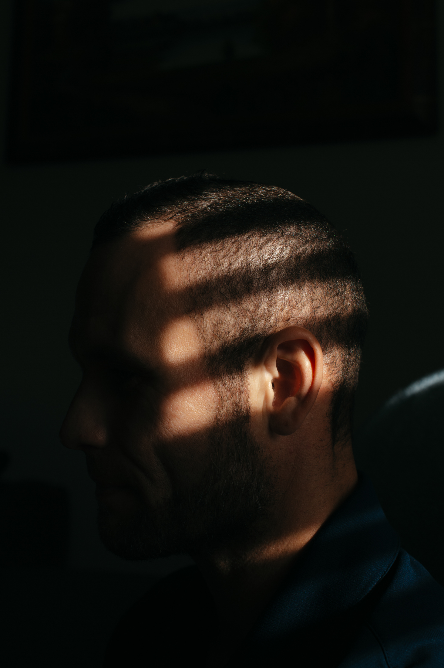A closeup of a man's head in a dark room, with lines of light from the window blinds illuminating parts of his head.