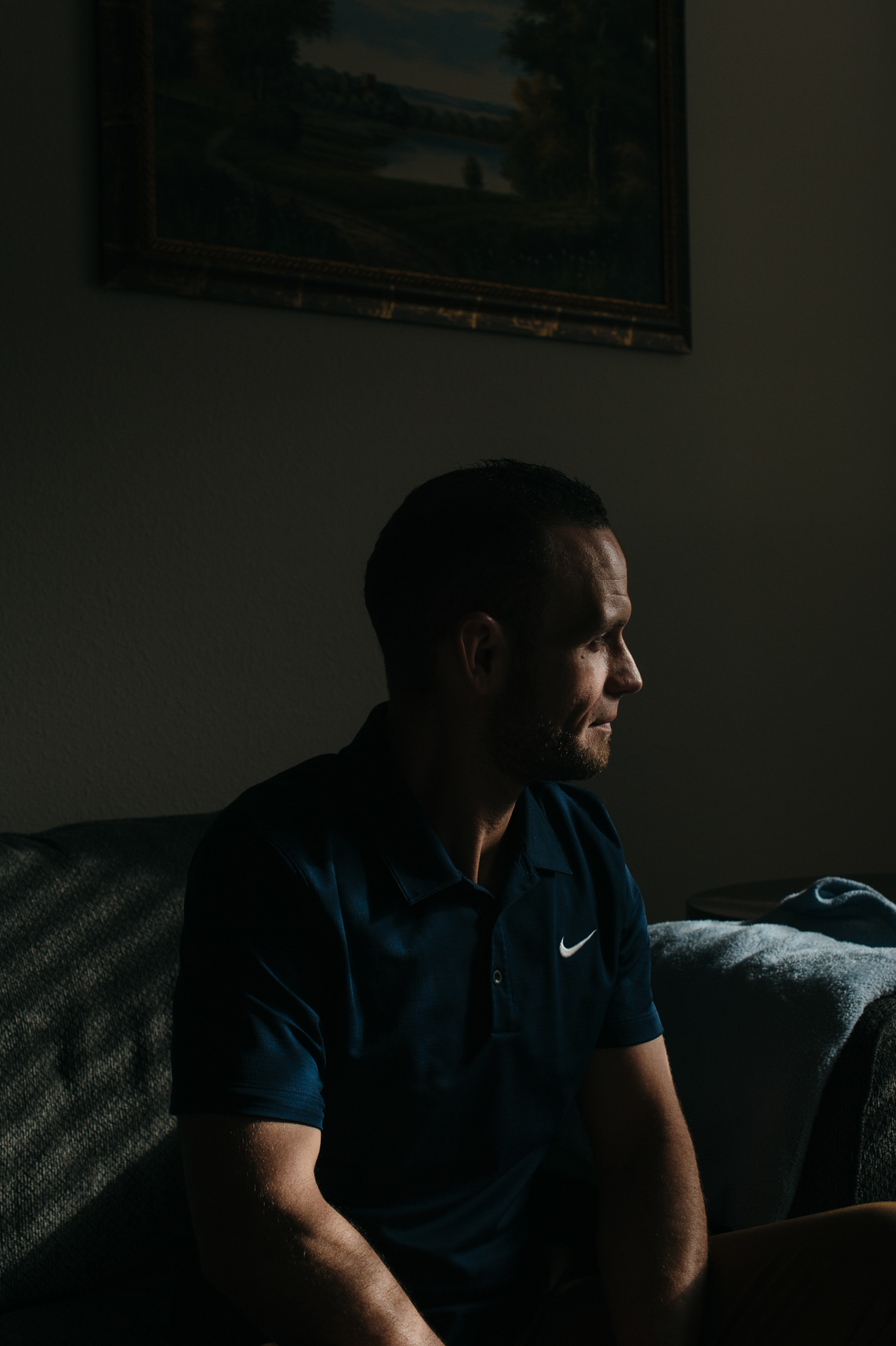 A man sits in a dark room, with his face slightly illuminated by a nearby window.
