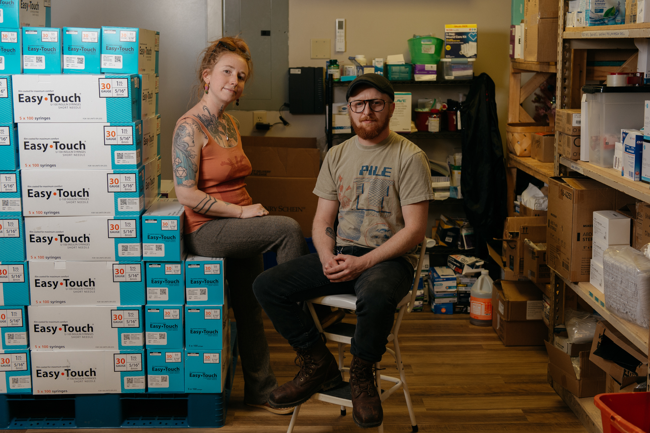 A woman sits on a pile of needle boxes. A man sits in a chair beside her.