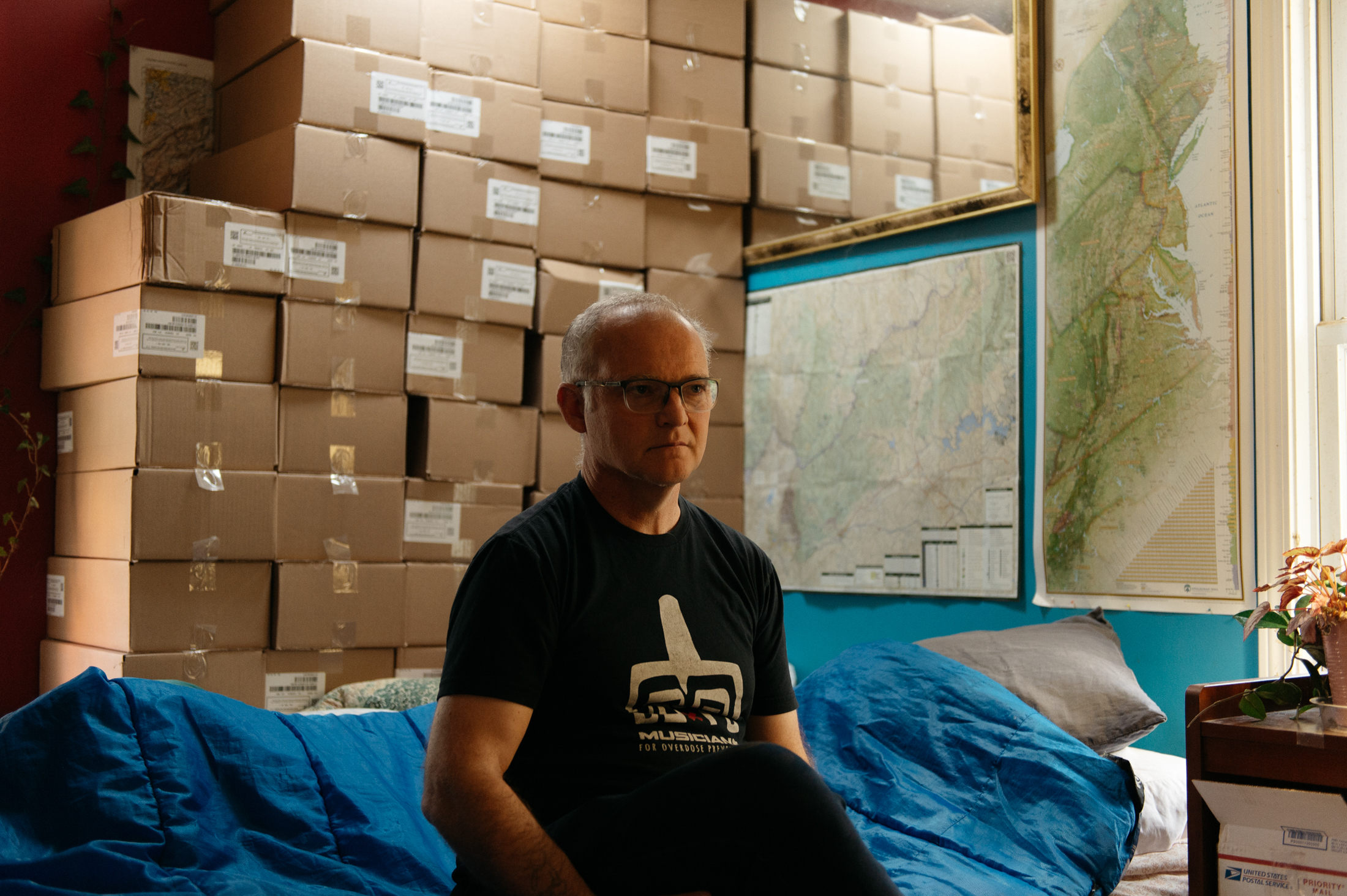 A man sits in front of a large stack of cardboard boxes containing Naloxone.
