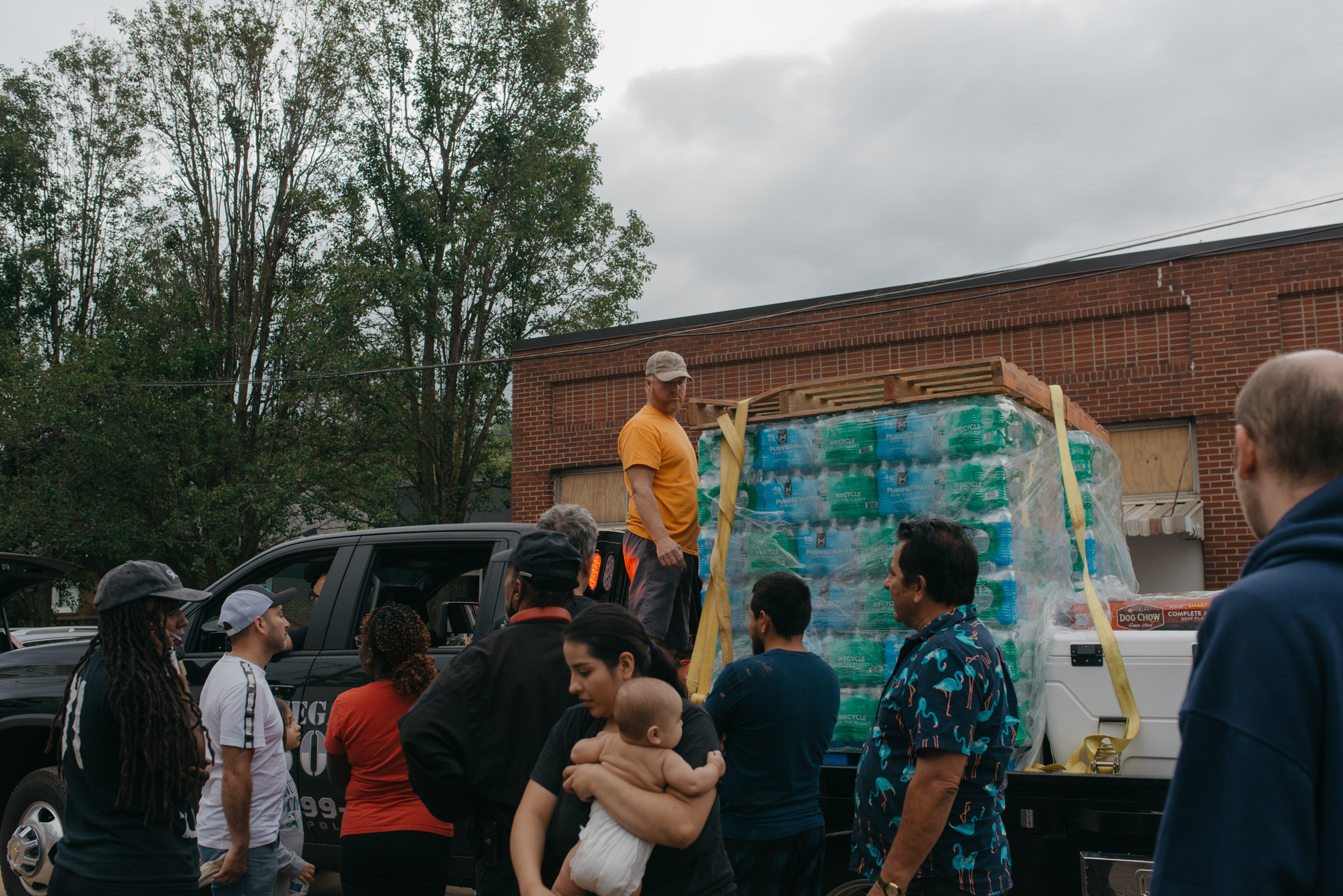 A man stands next to a palette of water bottles in the back of a pickup truck. A small crowd of people stands around the truck, waiting for supplies.