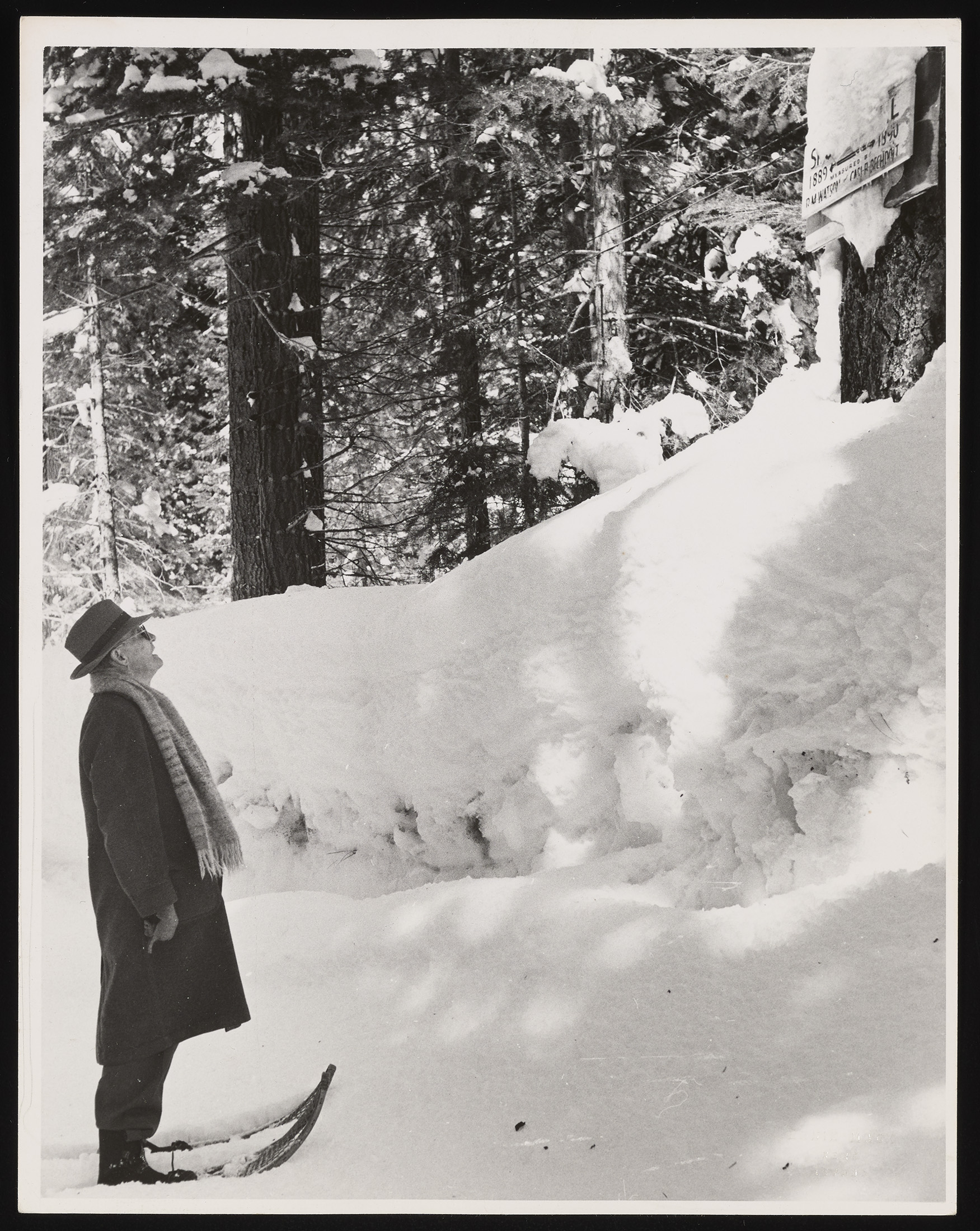 A man in a black and white photo stares up at a snow drift. He is wearing skis and a coat and hat