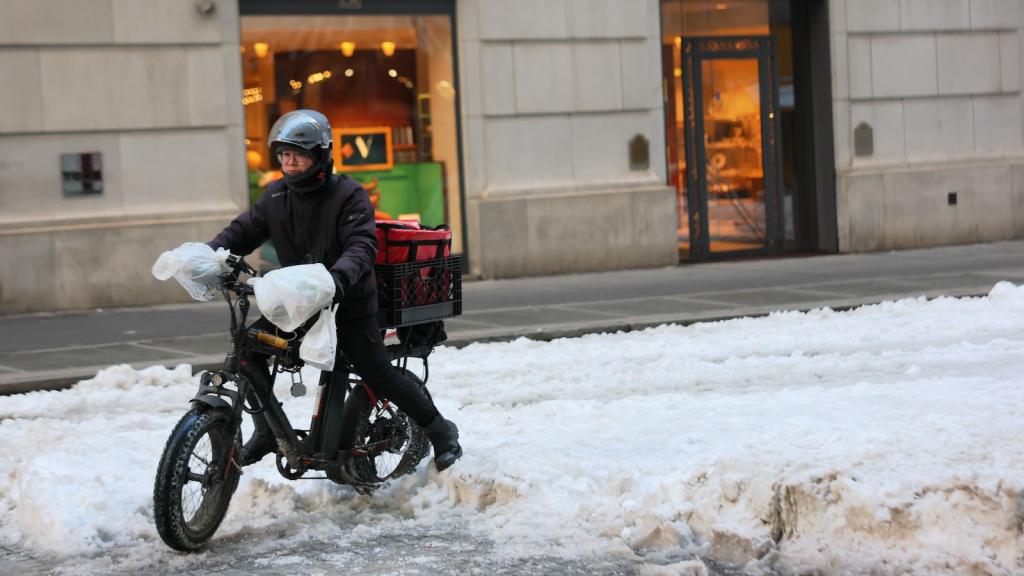 a delivery worker on an e-bike, wearing a bike helmet and a heavy winter jacket and black pants, rides through the streets of downtown Manhattan near the Stock Exchange, with several inches of visible snow on the ground