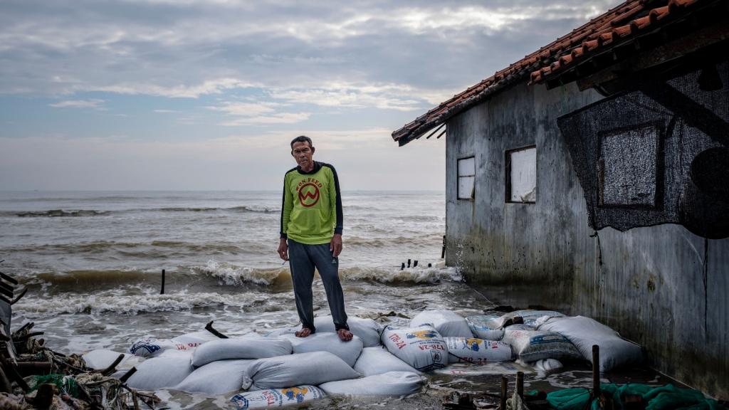 A man stands on sandbags next to a building as waves lap behind him