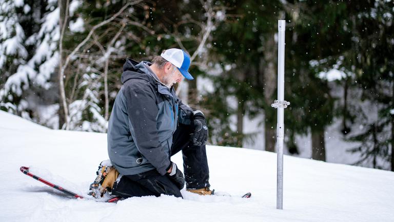Toby Rodgers, dressed in snowshoes and winter clothes, kneels in a snowy field, and looks at a long aluminum tube stuck into the snow.