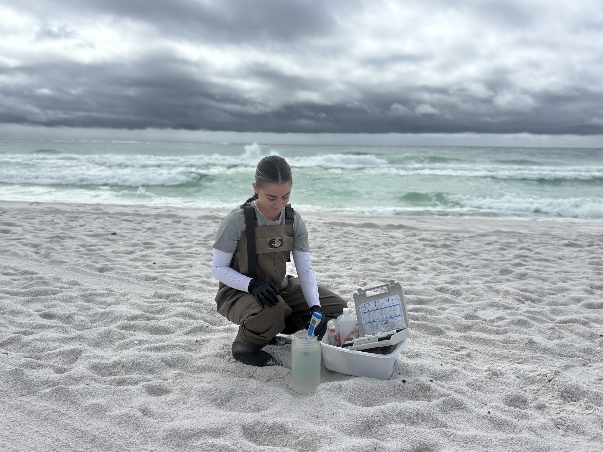 A woman kneels on a beach gathering samples of seawater
