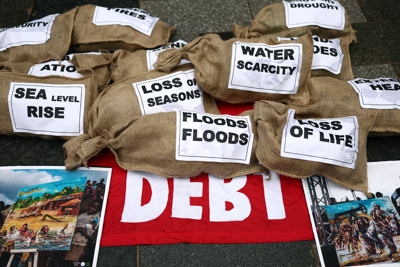 Photo of a protest with sacks put on top of a sign labeled "debt," naming water scarcity, floods, loss of life, sea level rise