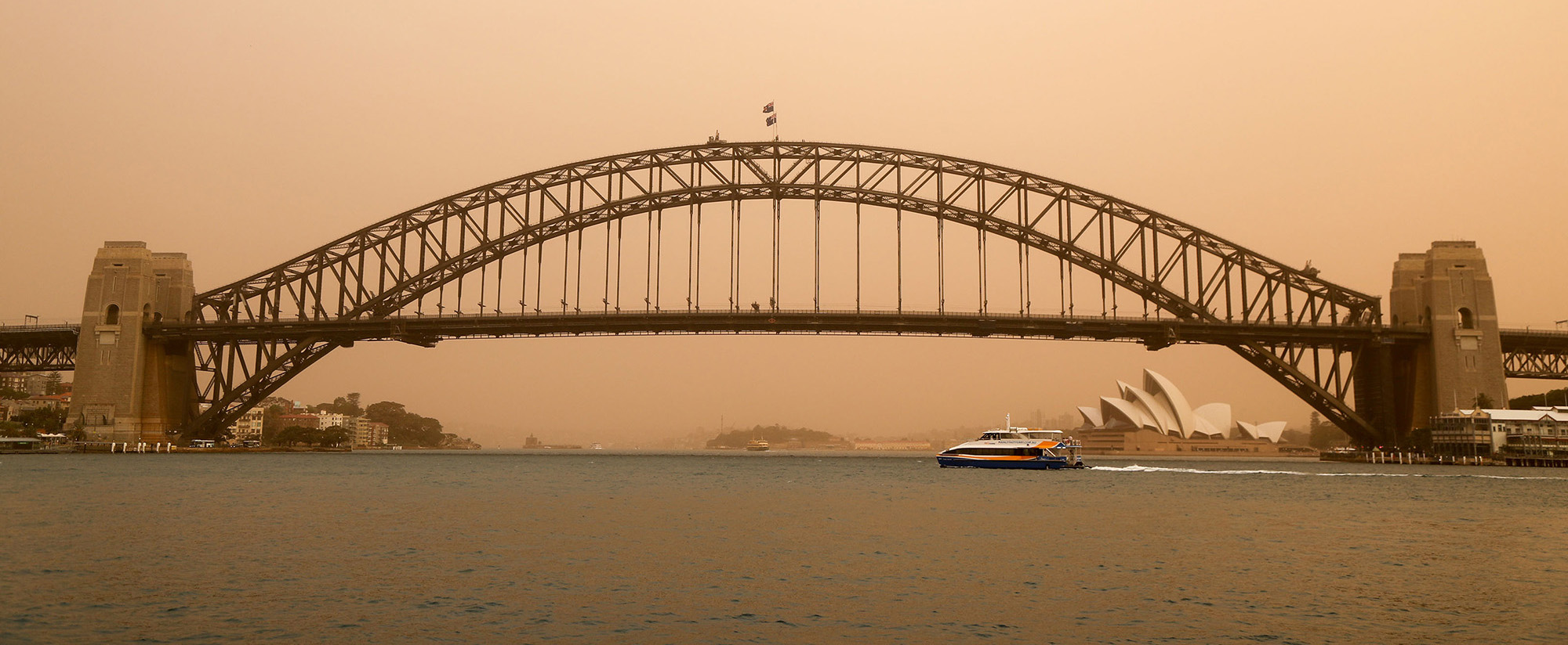 A photo of the Sydney Harbor Bridge in Australia, under an orange sky from wildfire smoke.