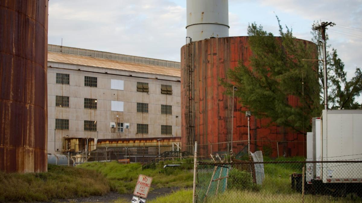 A rusting cylinder sits next to an industrial plant