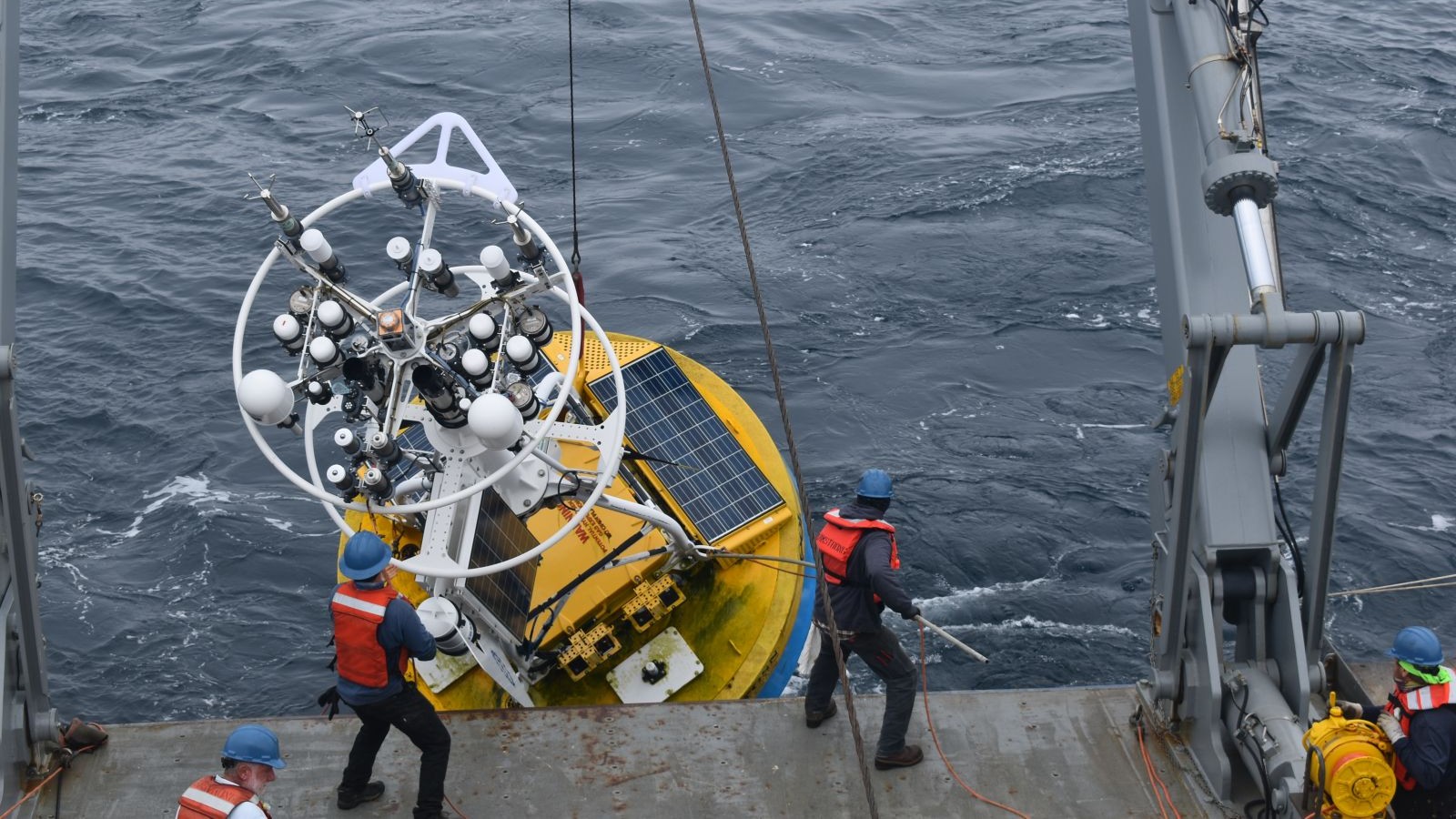 a large yellow instrument the size of a car goes into the ocean off of the side of a ship with crew in orange vests surrounding it