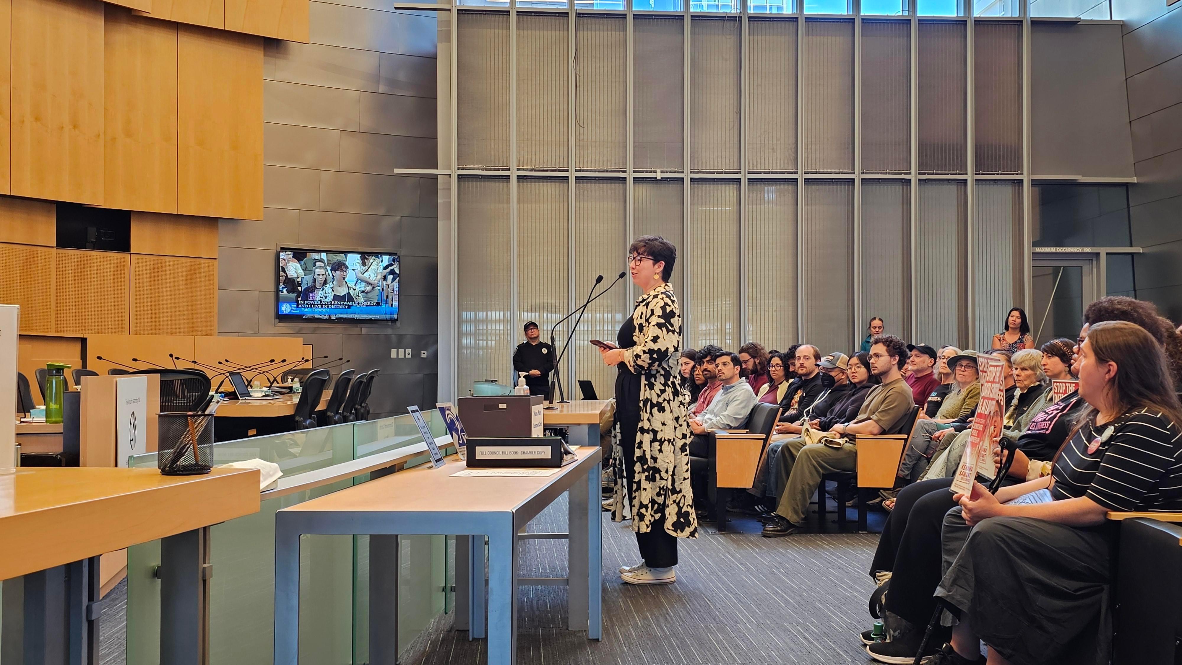Photo of a person speaking into a microphone in a room full of people