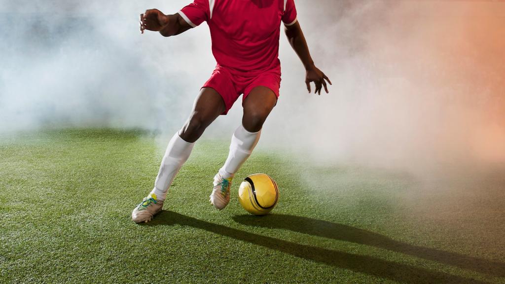 A soccer player kicks a yellow ball on turf with a haze of smoke around him
