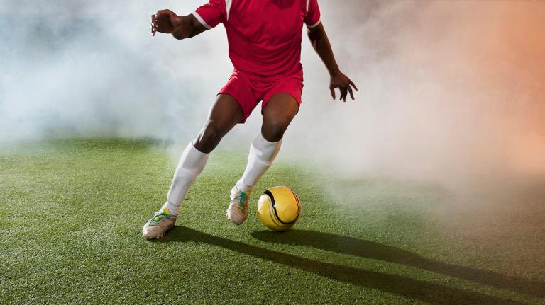 A soccer player kicks a yellow ball on turf with a haze of smoke around him